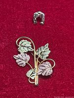 Two gold pins on red velvet background: a multi-colored leaf brooch and a small horseshoe pin.
