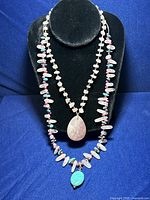 Two necklaces displayed on a black necklace stand. One necklace is predominantly rose quartz beads with a large teardrop-shaped rose quartz pendant. The other necklace includes irregular beads in turquoise, purple, and rose quartz with a turquoise pendant.