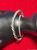 Side view of twisted cable bracelet and mesh link bracelet displayed on black stand with red background, showing texture and finish of both pieces.