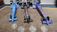 Three Bandameer Speedway blue and black dragster collector models positioned upright on carpet along with four NHRA 'Troops on the Track' collector coins placed in front of each dragster.