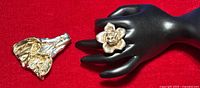 Photo of silver flower brooch and rose ring displayed on black mannequin hand over red background.