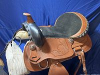 Full view of brown tooled leather saddle with black suede seat, silver-tone conchos and braided trim