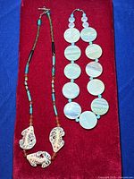 Full view of two necklaces on red background; jasper/agate disc necklace with blue spacers and carved fish pendant necklace with beads.