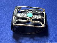 Front view of sterling silver belt buckle with leaf patterns and an oval turquoise stone center on black leather belt.