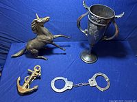Photo showing brass unicorn figurine, large metal trophy cup with two handles, brass seaman's anchor, and metal handcuffs on blue fabric background.