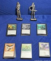 Two pewter warrior woman statues and six stacks of Magic The Gathering cards laid out on a blue cloth background. The statues depict warrior women in armor holding swords with distinct poses. The cards show varied artworks and colors indicating different card types.