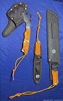 Three-piece set showing tactical axe with black nylon sheath and orange paracord handle, smaller knife with sheath and paracord lanyard, and additional fixed blade knife also sheathed.