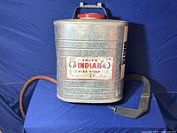Front view of vintage galvanized metal Smith Indian fire pump can on blue background showing red top and labeled front with attached hose and strap.
