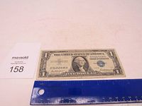 Front view of the 1957 Blue Seal Silver Certificate One Dollar bill with a blue ruler for size reference, showing overall good condition and details like the blue seal and serial number.