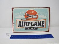 Front view of the rectangular metal airplane rides sign with a plane flying illustration and orange sun.