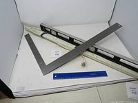 Full view of the lot showing the white electrical power strip, metal carpenter's square, black plastic level tool, and blue plastic ruler on a white surface.