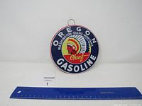 Full view of the Oregon Chief Gasoline metal round sign hanging against a white wall with a blue ruler placed below it for size reference.