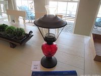 Full view of the red glass oil lamp showing the red glass font, black metal base and top, and clear glass chimney.