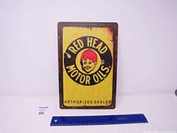 Front view of the Red Head Motor Oils metal sign showing the yellow background with circular black logo and red-headed boy illustration, with rust and wear marks.