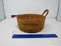 Front angle of the round woven basket with one handle upright and one folded, shown with a ruler for scale