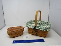 Photo showing two Longaberger baskets side by side on a white backdrop with a ruler for scale - one larger with handle and green leaf fabric liner, one smaller with floral liner.