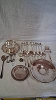 Overview of assorted silverplated dishes and utensils arranged on cloth background including spoons, forks, tongs, plates and decorative items.