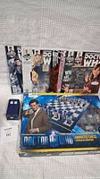 Photo showing seven Doctor Who comics aligned upright, a boxed Doctor Who animated chess game, and a small TARDIS-themed item.