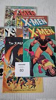 Top view of multiple Marvel The Uncanny X-Men comic books and an X-Men fan magazine arranged overlapping, showing key cover art and colorful designs.
