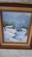 Framed original acrylic painting showing a snow-covered rural landscape with buildings and a stream under a cloudy sky.
