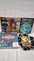 Star Trek trivia board game box, magazines, book about The Next Generation starship, fabric, and car booklet arranged on a bedspread.