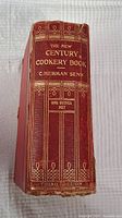 Side spine view showing the red half leather hardcover with gold gilt embossed text and decoration, titled 'The New Century Cookery Book C. Herman Senn, One Guinea Net, Third Edition'. The leather binding shows wear at edges.
