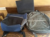 Photo showing the two backpacks and black portfolio folio arranged on a wooden bench.