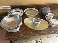 Photo showing assortment of kitchenware items on wooden sideboard including metal mixing bowl, cream and sugar set, tea pot, ceramic and serving bowls, Corning Ware casserole dish, and mortar and pestle.