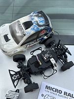 Micro rally car with white and blue painted body shell, exposed chassis showing four-wheel drive, battery compartment, and tires. Accompanying instruction manual visible in the background.