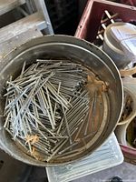 Metal container filled with a large quantity of small silver finish nails, placed in a crate with other miscellaneous hardware items.