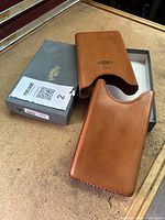 Two brown leather cigar holders displayed with branded grey Savinelli box on a wood surface. One holder inside the box, the other resting beside it.