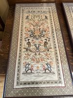 First embroidered wall hanging featuring symmetrical design with floral elements, human figures, and stylized animals framed with patterned fabric mat.