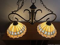 Two light Tiffany style stained glass pendant lamp with yellowish cream, white, and amber colors, bronze metal frame with decorative curved arms and hanging chains