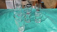 Six beer glasses with brewery logos including Muskoka, Samuel Adams, Black Oak, Left Field, Grey Matter, and Cameron's arranged on green tarp.