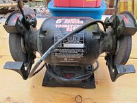 Front view of black 6 inch bench grinder showing motor housing with label and power switch, tool rests and protective eye shields on each grinding wheel side.