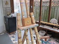Front view of wooden art easel showing paint stains and adjustable canvas holder with landscape of room background.