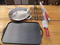 Photo showing all items on wooden surface including rectangular grilling pan, perforated round grilling pan, fish rack, grilling fork and spatula with Budweiser branding