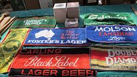 Photo of various branded bar towels laid out, showing logos from Tennent's Pilsner, Foster's Lager, Carling, Molson Canadian, and Double Diamond beers.