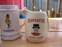 Photo showing two ceramic drinkware items: Planters Mr. Peanut mug and Beefeater Dry Gin water jug on wooden table.