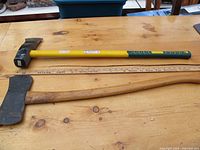 Photo of a vintage double-headed axe with a wooden handle and a yellow and green handled log splitter lying side by side on a wooden surface with a ruler for scale.