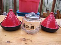Two Ethiopian style tagine pots with red conical lids and black bases alongside a clear glass cookie jar with metal lid labeled COOKIES, arranged on wooden surface.