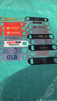 All 13 flat metal bottle openers laid out on a light blue surface showing brand logos and colors