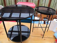 Two round black metal frame side tables with smoked black glass tops and lower shelves. One table is larger (24") and one smaller (21"). Both have gold accents on legs.