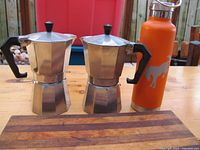 Front view of two aluminum Italian stovetop coffee makers, an orange Bronco water bottle, and a wooden cutting board on a wooden table outdoors.
