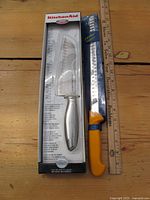 7 inch KitchenAid santoku knife in packaging with silver handle alongside 12 inch large kitchen knife with yellow handle and serrated blade next to ruler