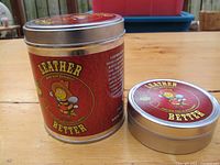 Two tins of Leather Better bee-based leather treatment on wooden surface, closed tins