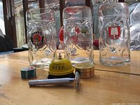 Four large 1-litre beer steins with embossed patterns and logos, yellow metal beer bell, and metal safety razor displayed on wooden surface.
