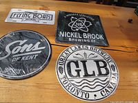 Four tin brewery signs laid out on wooden surface: rectangular Flying Boats, square Nickel Brook, round Sons of Kent (shrink-wrapped), round GLB.