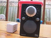Front view of black Tivoli Audio PAL portable radio on table with visible tuning dial, volume and power knobs, speaker grille and white AC power transformer connected.