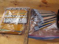 Photo showing two large plastic bags: one filled with unsharpened yellow HB pencils, the other with assorted painting brushes.